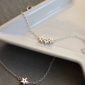 Two bracelets are shown draped over a star dish. One has 3 stars all the same size each with a different initial. The other bracelet in the foreground has one star with a single initial and hammered texture.