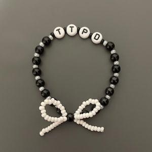 May include: Black and silver beaded bracelet with white beads spelling out the word "TTPD" and a white bow.
