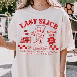 May include: White t-shirt with red text that reads "Last Slice of the Stephanie's Bachelorette Single Life Est. 2025". The shirt features a cartoon pizza slice with legs and arms.