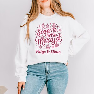 May include: White sweatshirt with a festive design in burgundy. The design includes the text "Soon To Be Merry'd" with decorative ornaments and the names "Paige & Ethan" underneath. The sweatshirt is paired with blue jeans.