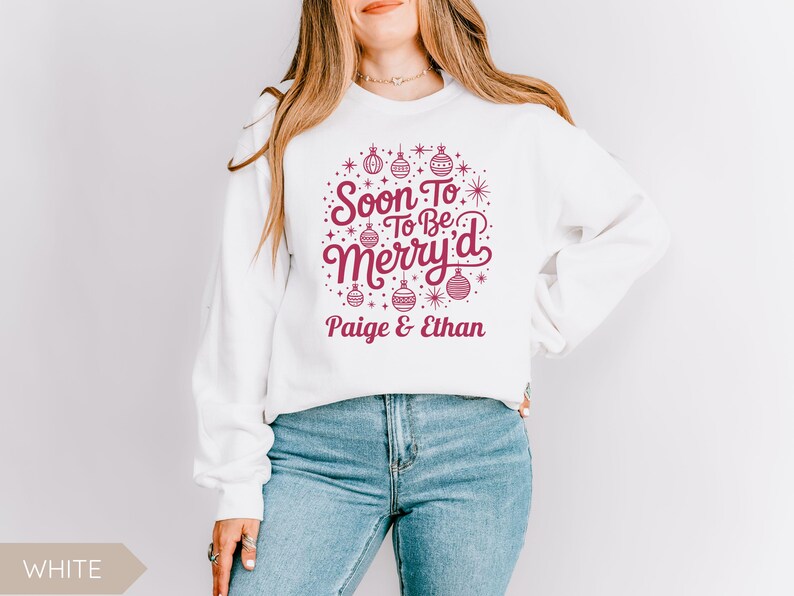 May include: White sweatshirt with a festive design in burgundy. The design includes the text "Soon To Be Merry'd" with decorative ornaments and the names "Paige & Ethan" underneath. The sweatshirt is paired with blue jeans.