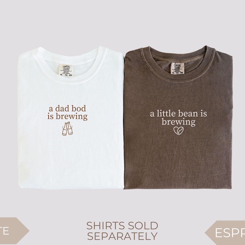 Two folded Comfort Colors t-shirts displayed side by side on a light background. The white shirt features the phrase “a dad bod is brewing” in brown text with a small illustration of two clinking bottles. The espresso brown shirt shows the phrase “a little bean is brewing” in white text with a tiny heart bean graphic. Matching pregnancy announcement shirts for expecting parents.