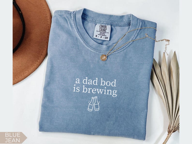 K&ouml;nnte beinhalten: Blaues Jeans-T-Shirt mit dem wei&szlig;en Schriftzug "a dad bod is brewing". Darunter befinden sich zwei wei&szlig;e Flaschenillustrationen. Eine goldene Halskette und ein brauner Hut sind ebenfalls zu sehen.