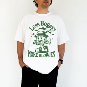 May include: White t-shirt with a green graphic of a cartoon golf ball wearing a hat and holding a golf club. The text "Less Bogeys More Blowies" is printed on the shirt.