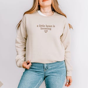 K&ouml;nnte beinhalten: Sandfarbenes Sweatshirt mit dem Text "a little bean is brewing" in brauner Schrift und einem Kaffeebohnen-Herz-Motiv. Das Sweatshirt wird mit einer blauen Jeans kombiniert.