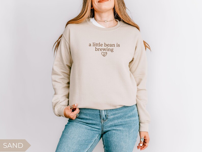 K&ouml;nnte beinhalten: Sandfarbenes Sweatshirt mit dem Text "a little bean is brewing" in brauner Schrift und einem Kaffeebohnen-Herz-Motiv. Das Sweatshirt wird mit einer blauen Jeans kombiniert.