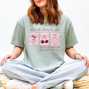 May include: A pale green t-shirt featuring the phrase "She's The Cherry On Top" and three illustrations. The illustrations include a cocktail, cherries, and a disco ball. The text "Olivia's Bachelorette Nashville, TN / 2026" is also printed on the shirt.