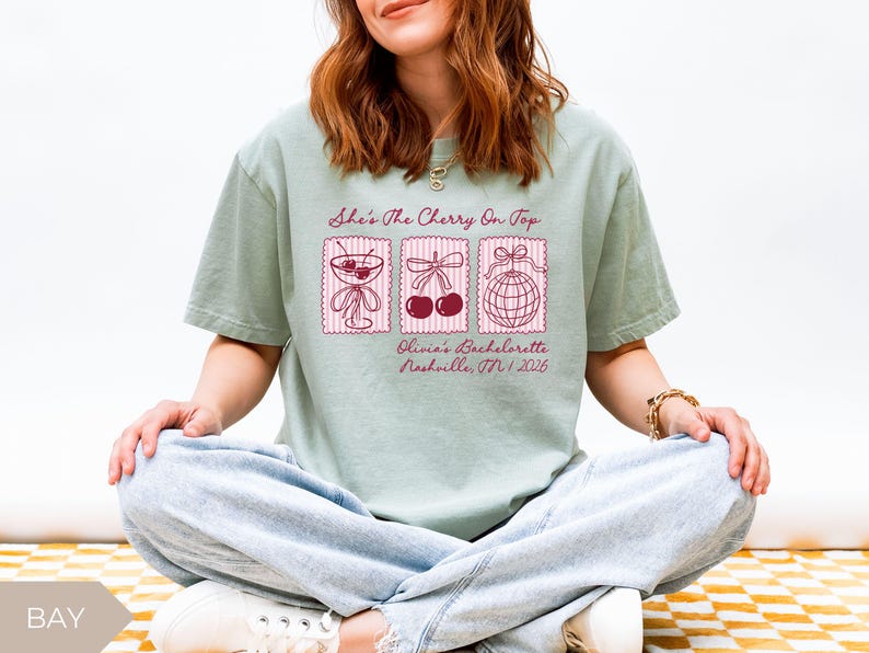 May include: A pale green t-shirt featuring the phrase "She's The Cherry On Top" and three illustrations. The illustrations include a cocktail, cherries, and a disco ball. The text "Olivia's Bachelorette Nashville, TN / 2026" is also printed on the shirt.