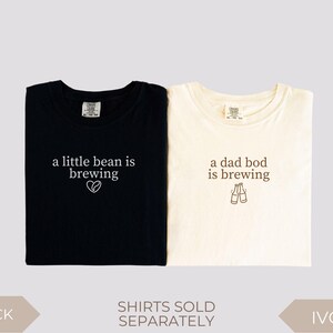 K&ouml;nnte beinhalten: Zwei gefaltete T-Shirts, eines in Schwarz und eines in Elfenbein, mit Text. Das schwarze Shirt liest "a little bean is brewing" mit einem Herzsymbol. Das elfenbeinfarbene Shirt sagt "a dad bod is brewing" mit zwei Bierflaschen-Illustrationen. Shirts werden separat verkauft.