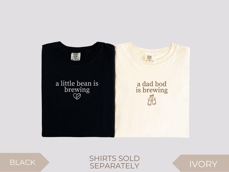 K&ouml;nnte beinhalten: Zwei gefaltete T-Shirts, eines in Schwarz und eines in Elfenbein, mit Text. Das schwarze Shirt liest "a little bean is brewing" mit einem Herzsymbol. Das elfenbeinfarbene Shirt sagt "a dad bod is brewing" mit zwei Bierflaschen-Illustrationen. Shirts werden separat verkauft.