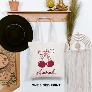 May include: A cream tote bag featuring a burgundy cherry design with a bow and the name "Sarah". The bag is displayed on a wall with a black hat, a shelf, and a decorative wall hanging.