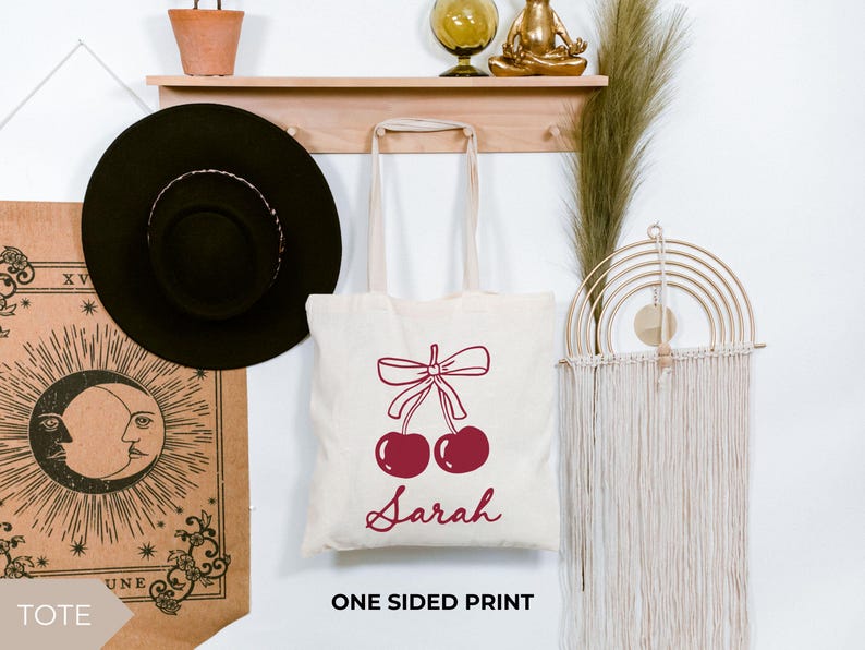 May include: A cream tote bag featuring a burgundy cherry design with a bow and the name "Sarah". The bag is displayed on a wall with a black hat, a shelf, and a decorative wall hanging.