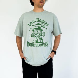 May include: A light blue t-shirt with a green cartoon golf ball character wearing a hat and holding a golf club. The text on the shirt reads "Less Bogeys More Blowies".