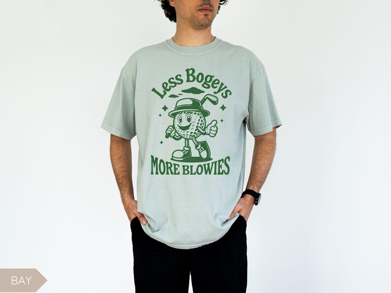 May include: A light blue t-shirt with a green cartoon golf ball character wearing a hat and holding a golf club. The text on the shirt reads "Less Bogeys More Blowies".