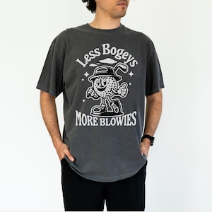 May include: A dark gray t-shirt with a white graphic of a cartoon golf ball character holding a golf club. The text "Less Bogeys More Blowies" is printed on the shirt.