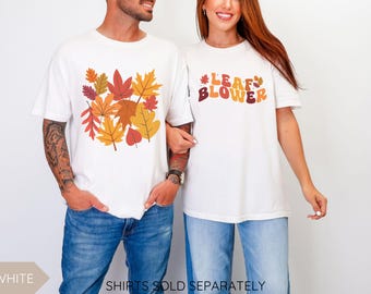 May include: White t-shirts with fall-themed graphics. One shirt features a collection of colorful autumn leaves, while the other displays the words "LEAF BLOWER" in a retro font. Both shirts are paired with blue jeans.