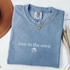 May include: A blue jean Comfort Colors t-shirt with the phrase "bun in the oven" printed in white. A gold necklace with a pendant rests on the shirt. A brown hat and dried plant are in the background.