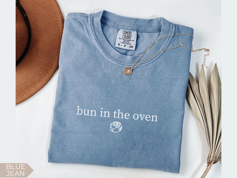 May include: A blue jean Comfort Colors t-shirt with the phrase "bun in the oven" printed in white. A gold necklace with a pendant rests on the shirt. A brown hat and dried plant are in the background.