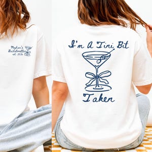 May include: White t-shirt with a navy blue martini glass graphic and the text "I'm a Tini Bit Taken" on the back. The front of the shirt has the text "Meghan's Bachelorette est. 2026" and a martini glass graphic.