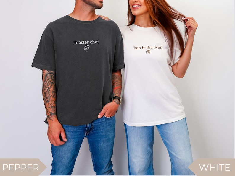 May include: Two short-sleeved t-shirts, one dark gray and one white. The dark gray shirt has "master chef" printed on it with a chef's hat graphic. The white shirt says "bun in the oven" with a bun graphic. Both shirts are paired with blue jeans.