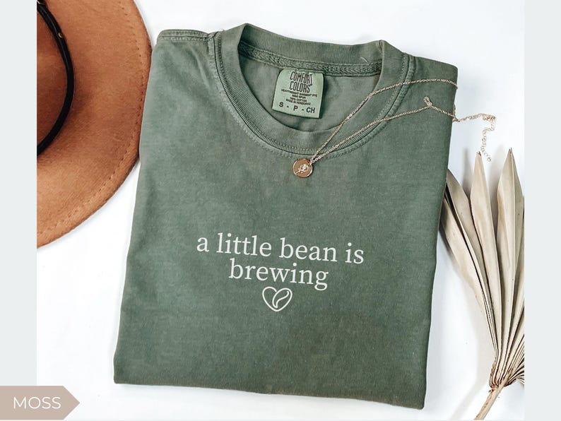 K&ouml;nnte beinhalten: Moosgr&uuml;nes T-Shirt mit dem Schriftzug "a little bean is brewing" und einer Kaffeebohnen-Herz-Grafik. Eine goldene Halskette und ein brauner Hut sind ebenfalls im Bild. Das Shirt ist von der Marke Comfort Colors.