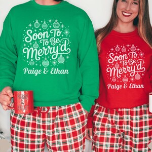 May include: Green and red Christmas-themed sweatshirts with the text "Soon To Be Merry'd" and the names "Paige & Ethan". The sweatshirts are paired with red and white plaid pajama pants. One person holds a red mug that says "Merry & Bright", and the other holds a white mug.