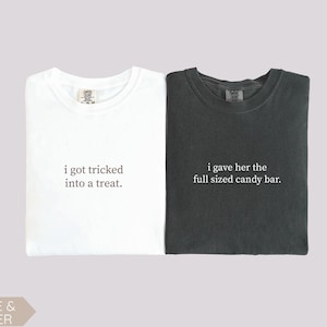 May include: Two t-shirts, one white and one dark gray, with text. The white shirt says "i got tricked into a treat." The dark gray shirt says "i gave her the full sized candy bar."