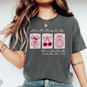 May include: Dark grey t-shirt with the text "She's The Cherry On Top" and three pink postage stamp designs. The stamps feature a cocktail, cherries, and a disco ball. Below the stamps is "Olivia's Bachelorette Nashville, TN / 2026".