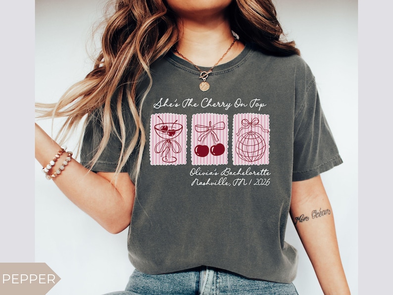 May include: Dark grey t-shirt with the text "She's The Cherry On Top" and three pink postage stamp designs. The stamps feature a cocktail, cherries, and a disco ball. Below the stamps is "Olivia's Bachelorette Nashville, TN / 2026".