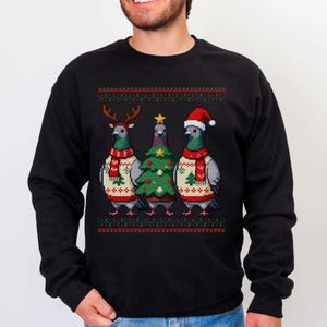 May include: Black sweatshirt featuring three pigeons dressed for Christmas. One pigeon wears reindeer antlers, another a Christmas tree, and the third a Santa hat. Each bird has a festive sweater and scarf.