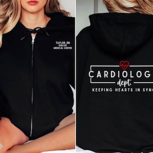May include: Black zip-up hoodie with a white graphic that says "Cardiology dept. Keeping hearts in sync" with a red heart and EKG line. The hoodie also has white text that says "Taylor, RN Dallas Medical Center".