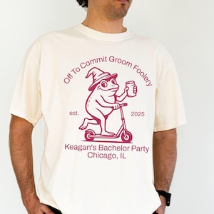 May include: A cream-coloured t-shirt with a maroon graphic of a frog on a scooter, wearing a wizard hat, and holding a mug. Text reads "Off To Commit Groom Foolery" and "Keagan's Bachelor Party Chicago, IL".