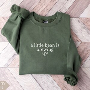 K&ouml;nnte beinhalten: Milit&auml;rgr&uuml;nes Sweatshirt mit dem wei&szlig;en Text "a little bean is brewing" und einem Kaffeebohnen-Herz-Motiv. Das lang&auml;rmlige Oberteil mit Rundhalsausschnitt ist aus weichem Material gefertigt.