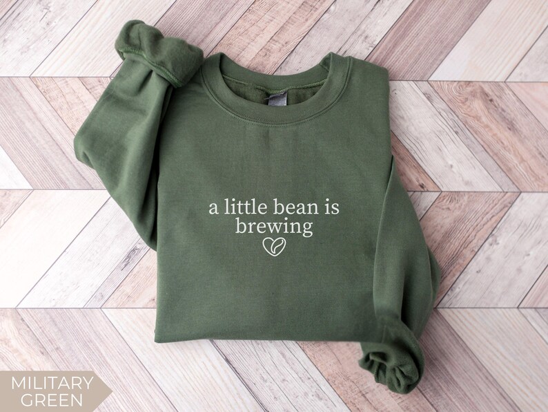 K&ouml;nnte beinhalten: Milit&auml;rgr&uuml;nes Sweatshirt mit dem wei&szlig;en Text "a little bean is brewing" und einem Kaffeebohnen-Herz-Motiv. Das lang&auml;rmlige Oberteil mit Rundhalsausschnitt ist aus weichem Material gefertigt.