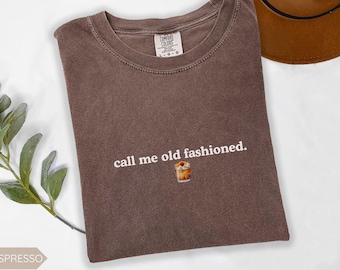 May include: Espresso-colored t-shirt with the phrase "call me old fashioned." in white script. A small graphic of an Old Fashioned cocktail is below the text. The shirt is folded and has a tag at the neckline.