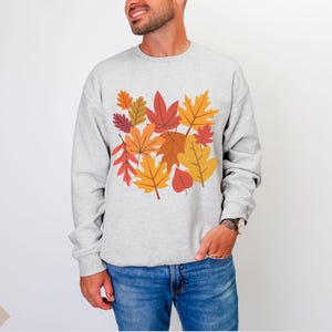 May include: A light gray sweatshirt featuring a colorful autumn leaves design. The leaves are in shades of red, orange, and yellow. The sweatshirt is a crew neck style and is made of a soft material.