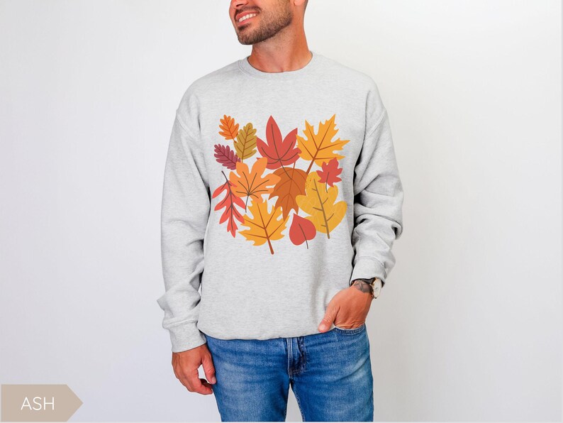 May include: A light gray sweatshirt featuring a colorful autumn leaves design. The leaves are in shades of red, orange, and yellow. The sweatshirt is a crew neck style and is made of a soft material.