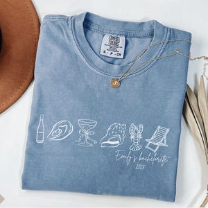 May include: A blue jean Comfort Colors t-shirt featuring a white outline graphic of a bottle, oyster, cocktail, seashell, lobster, and deckchair. The text reads "Emily's bachelorette 2025". A gold necklace and a brown hat are also visible.