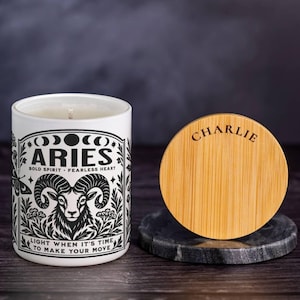 May include: A white cylindrical candle jar featuring a black graphic print of a ram, the text ARIES, BOLD SPIRIT, FEARLESS HEART, and LIGHT WHEN IT'S TIME TO MAKE YOUR MOVE. Beside it sits a round bamboo lid engraved with the name CHARLIE.