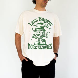 May include: A white t-shirt with a green graphic of a cartoon golf ball character holding a golf club. The text "Less Bogeys More Blowies" is printed on the shirt.