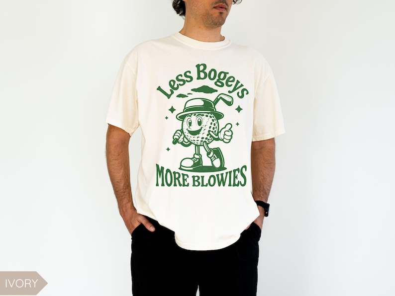 May include: A white t-shirt with a green graphic of a cartoon golf ball character holding a golf club. The text "Less Bogeys More Blowies" is printed on the shirt.