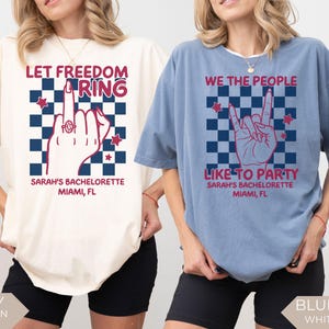Two women wearing oversized Comfort Colors t shirts in ivory and blue jean colors with patriotic graphic designs for a 4th of July bachelorette party. One shirt reads “Let Freedom Ring” with a hand graphic and custom name and location text. The other shirt reads “We the People Like to Party” with a rock hand gesture design. Styled for a summer girls trip or patriotic bach celebration.