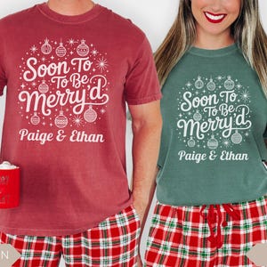 Two people wearing matching “Soon To Be Merry’d” Christmas shirts personalized with the names Paige and Ethan. The man wears a crimson red tee and the woman wears a light green tee, both paired with red and green plaid pajama pants and holding holiday mugs. The design features white ornaments and festive stars, perfect for engaged couples celebrating Christmas.