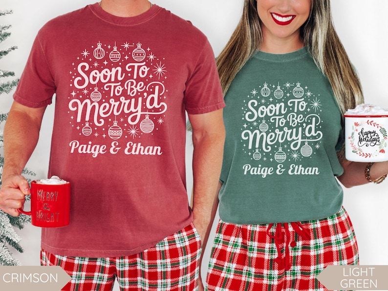 Two people wearing matching “Soon To Be Merry’d” Christmas shirts personalized with the names Paige and Ethan. The man wears a crimson red tee and the woman wears a light green tee, both paired with red and green plaid pajama pants and holding holiday mugs. The design features white ornaments and festive stars, perfect for engaged couples celebrating Christmas.