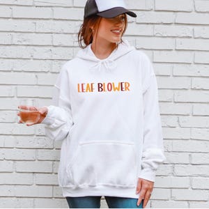 May include: White hooded sweatshirt with the text "LEAF BLOWER" in orange, yellow, and brown. The sweatshirt has a front pocket and a drawstring hood. The person in the image is wearing a black and white trucker hat.