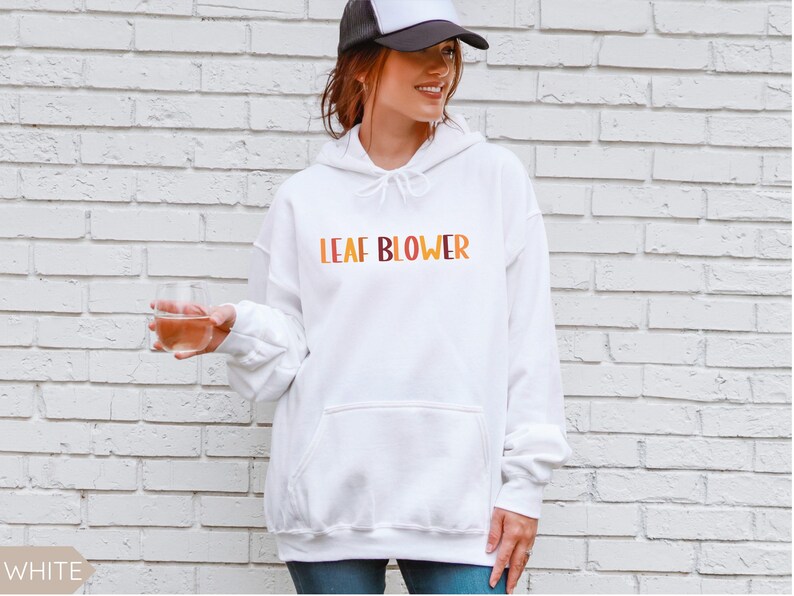 May include: White hooded sweatshirt with the text "LEAF BLOWER" in orange, yellow, and brown. The sweatshirt has a front pocket and a drawstring hood. The person in the image is wearing a black and white trucker hat.