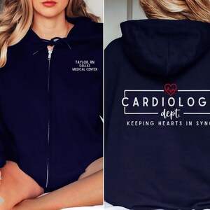 May include: Navy blue zip-up hoodie with a white graphic that says "Cardiology dept. Keeping hearts in sync" with a red heart and EKG line. The left side of the hoodie has white text that says "Taylor, RN Dallas Medical Center".