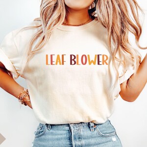 May include: Ivory-colored t-shirt with the words "LEAF BLOWER" in orange and brown lettering. The shirt has rolled-up sleeves and is paired with blue denim jeans. The overall style is casual and relaxed.