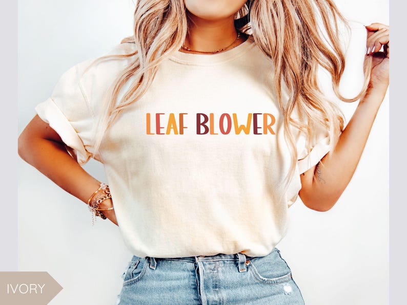 May include: Ivory-colored t-shirt with the words "LEAF BLOWER" in orange and brown lettering. The shirt has rolled-up sleeves and is paired with blue denim jeans. The overall style is casual and relaxed.