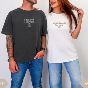K&ouml;nnte beinhalten: Zwei passende T-Shirts, eines in Dunkelgrau und eines in Wei&szlig;, mit Text. Das dunkelgraue Shirt tr&auml;gt die Aufschrift "a dad bod is brewing" mit einer Bierglas-Grafik. Das wei&szlig;e Shirt tr&auml;gt die Aufschrift "a little bean is brewing" mit einer Herzgrafik. Beide Shirts werden mit Jeans getragen.